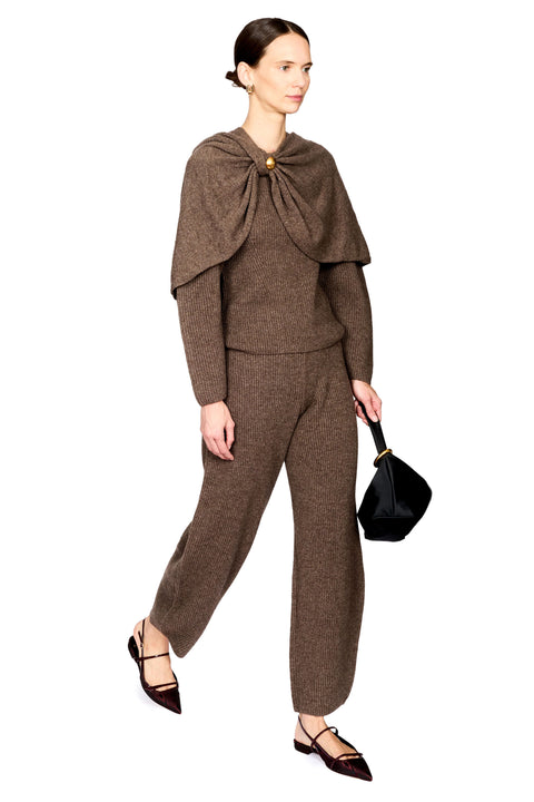 A model wearing a brown knit matching set consisting of a long-sleeve top with a draped collar and broch, and matching trousers, styled with dark pointed flats and a black handbag against a white background.