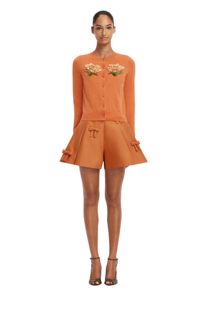 A model wearing a matching burnt orange two-piece set consisting of a long-sleeve button-front top with floral embroidery on the chest and matching flared shorts with side pockets, styled with brown strappy heels against a plain white background.
