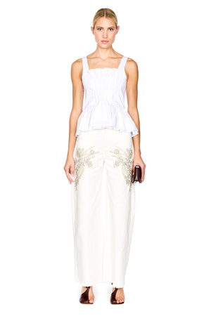 A woman modeling a white peplum top and white wide-leg trousers with gold floral embroidery, holding a small dark clutch against a plain white background.