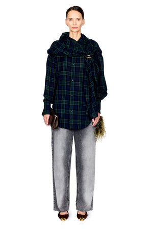 A model stands against a white studio background wearing a dark green and black plaid oversized button-up shirt with dramatic draped shoulders, light gray wide-leg jeans, and brown pointed shoes.