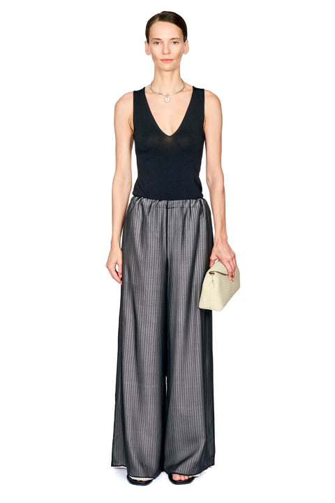 A model stands against a plain white background wearing a sleeveless black V-neck top and wide-leg charcoal gray pinstriped trousers, holding a small beige clutch.