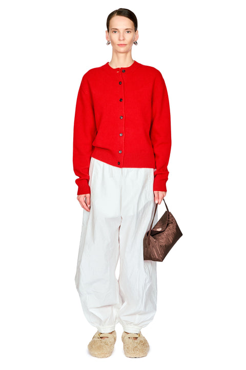 A model stands against a plain white background wearing a bright red button-front cardigan, loose white pants, beige slippers, and holding a brown textured handbag.