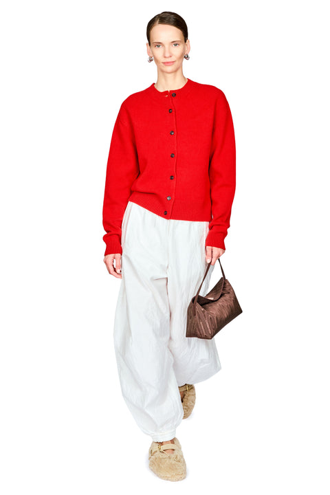 A model wearing a vibrant red button-up knit cardigan, loose white trousers, beige slippers, and holding a brown textured handbag against a plain white background.