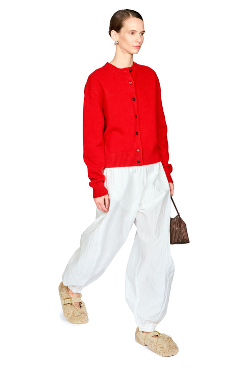 A model wearing a bright red knit cardigan, loose white trousers, beige knit loafers, and holding a small brown handbag against a plain white background.