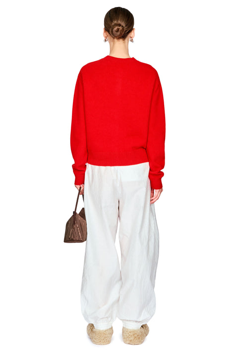 A model shown from the back wearing a bright red crewneck sweater, white wide-leg trousers, and beige woven slippers, holding a small brown handbag against a plain white background.