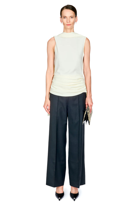 A model stands against a plain white background wearing a sleeveless white mock-neck top, dark black wide-leg trousers with white waist ruching, and black pointed heels, while holding a small patterned clutch.