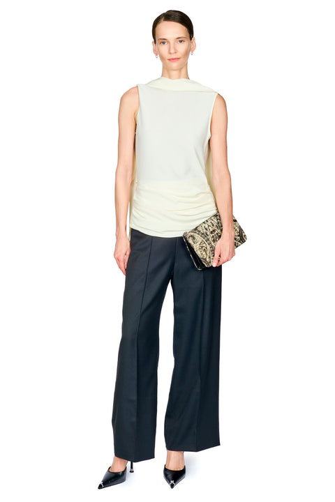 A model stands against a plain white background wearing a sleeveless white mock-neck top, dark black wide-leg trousers with white waist ruching, and black pointed heels, while holding a small patterned clutch.