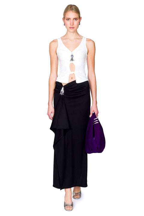 Woman wearing a white cutout top with a crystal pendant and black skirt clutching her purple bag.