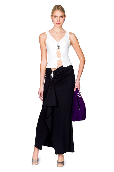 Woman wearing a white cutout top with a crystal pendant and black skirt with a purple bag on a white background