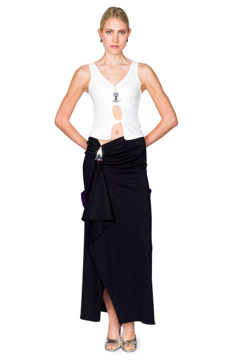 Woman wearing a white cutout top with a crystal pendant and black skirt with a purple bag on a white background with her hands in back.