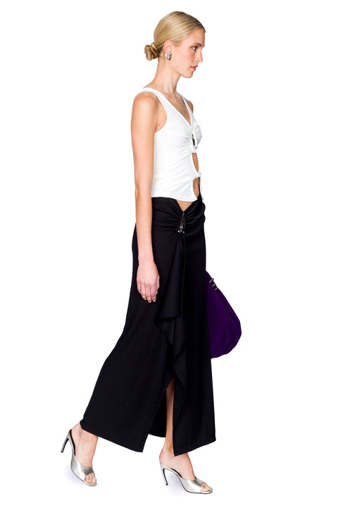 Woman wearing a white cutout top with a crystal pendant and black skirt with a purple bag on a white background from the side view.