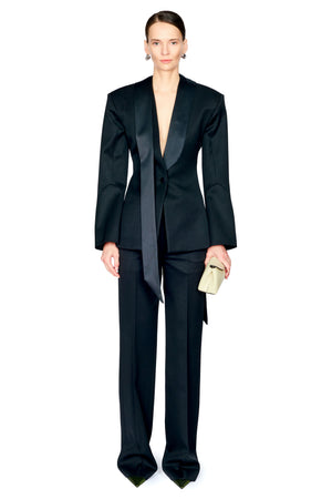 A model wearing a black satin and matte black two-piece suit with a deep V-neckline, wide lapels, and matching trousers, holding a small beige clutch against a plain white background.