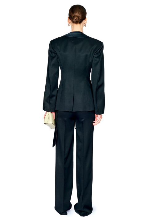 A model shown from the back wearing a black tailored suit, featuring a structured blazer with peak lapels and matching wide-leg trousers, holding a small light-colored clutch against a plain white background.