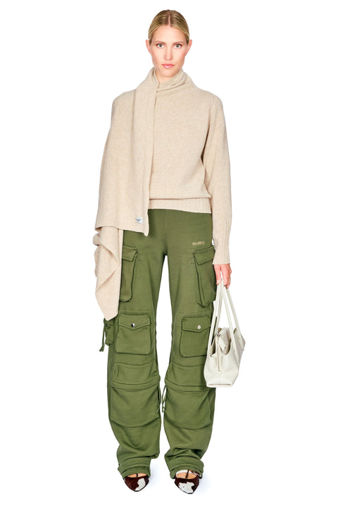 A woman wearing an oversized beige knit wrap sweater, olive green cargo pants, and holding a light beige tote bag.
