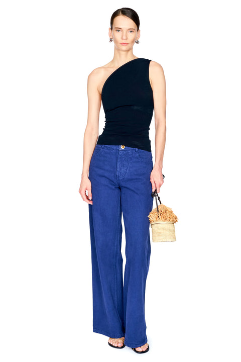 A model stands against a white background wearing a black one-shoulder top, blue wide-leg jeans, and holding a small beige woven handbag.