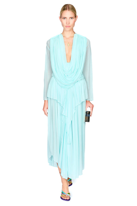 A model wearing a light aqua blue pleated long-sleeve top and maxi skirt set with a draped V-neckline, paired with colorful flat sandals and holding a black clutch, set against a plain white background.