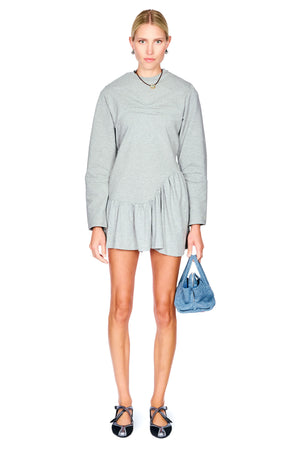 A woman modeling a light gray long-sleeve mini dress with a gathered waist, paired with black strappy heels and holding a blue handbag, against a plain white background.