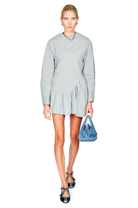 A model wearing a light gray long-sleeve mini dress with a ruffled hem, holding a blue denim handbag, and wearing black strappy heels against a plain white background.