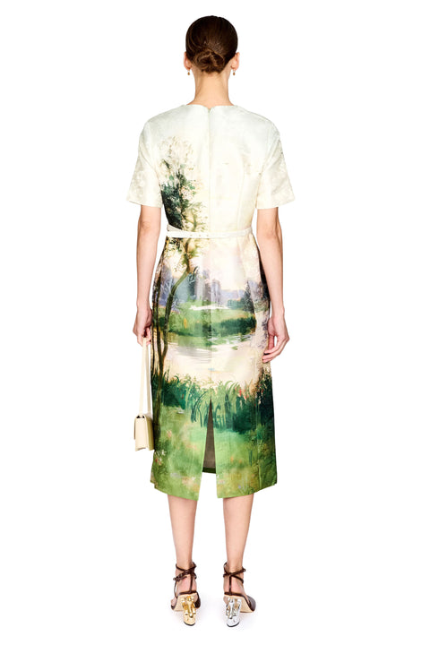 A model shown from the back wearing a short-sleeved midi dress with a scenic landscape print in green, blue, and cream tones. The dress features a tie waist and a front slit, paired with brown strappy heels and a small beige handbag against a plain white background.