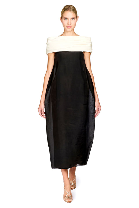 A woman modeling a black midi dress with an off-the-shoulder design featuring a contrasting cream-colored top band, set against a plain white background.