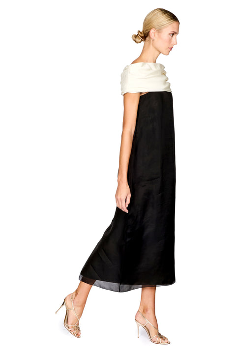 A woman modeling a black midi dress with a contrasting cream ruffled off-the-shoulder neckline, paired with beige high-heeled sandals, shown from a side profile against a white background.