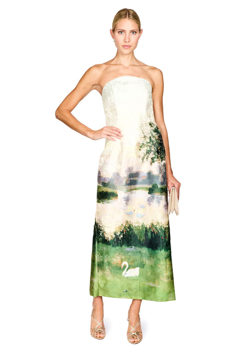 A woman modeling a strapless maxi dress with a white lace bodice and a green landscape print featuring a lake, trees, and a swan, set against a plain white background.