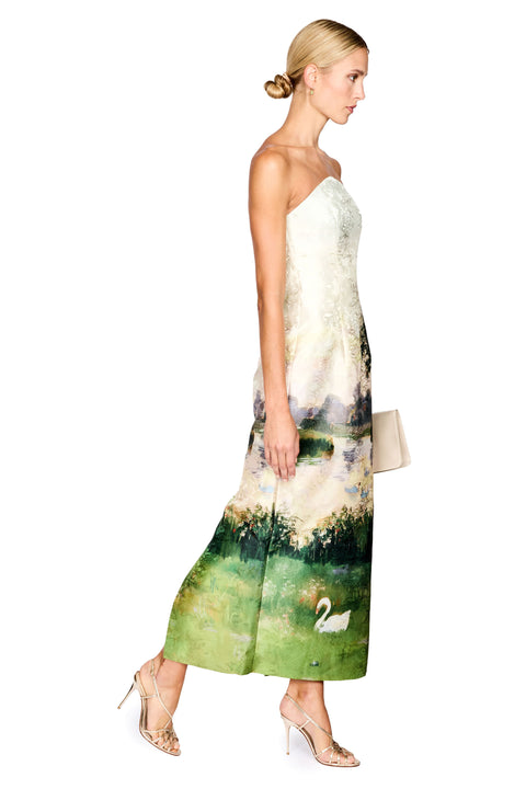 A woman modeling a strapless maxi dress with a scenic landscape print featuring green fields, trees, and a white swan, shown against a plain white background.