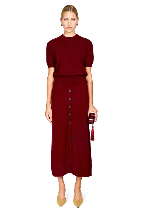 A woman modeling a matching burgundy knit short-sleeve top and midi skirt set with a button-front skirt, paired with beige heels and holding a small burgundy tassel bag, on a plain white background.