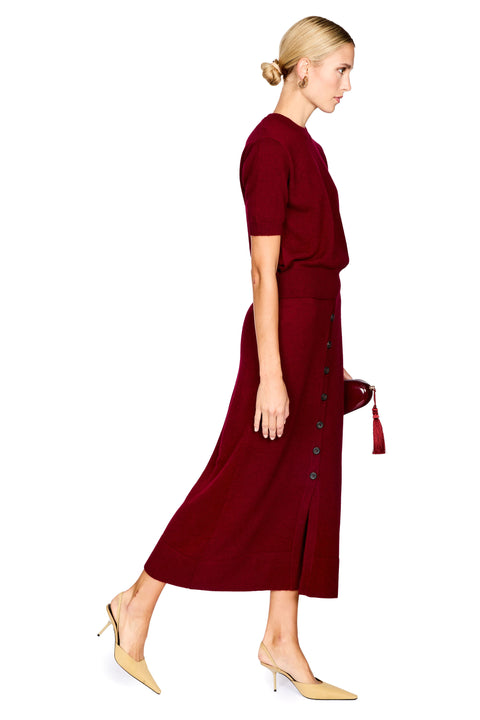 A woman in profile wearing a deep burgundy short-sleeve midi dress with a buttoned side detail, paired with beige pointed-toe heels, against a plain white background.