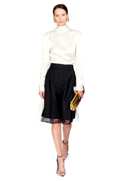 A model wearing a white long-sleeve mock-neck satin top, a black high-waisted pleated skirt with sheer black tulle hem, holding a gold clutch, and wearing black strappy high heels against a solid white background.