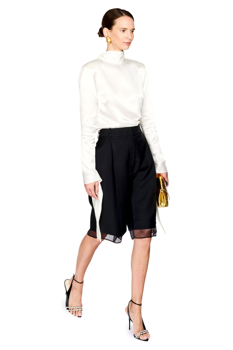 A model wearing a white long-sleeve mock neck top, black high-waisted shorts with sheer side panels, and black strappy high heels, holding a small gold handbag against a solid white background.