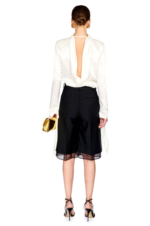 A model shown from the back wearing a white long-sleeve blouse with a deep open back, black high-waisted cropped trousers with a sheer black hem, and black strappy heels, holding a small gold handbag against a plain white background.