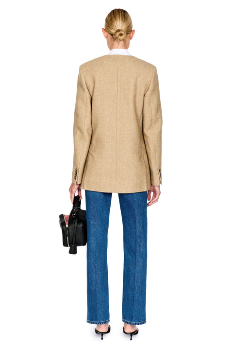 A woman shown from the back wearing a beige blazer, blue jeans, and black heels, holding a black handbag against a plain white background.
