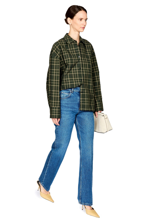 A woman modeling an oversized dark green and black plaid button-up shirt, paired with blue jeans, beige pointed-toe flats, and holding a small cream handbag against a plain white background.