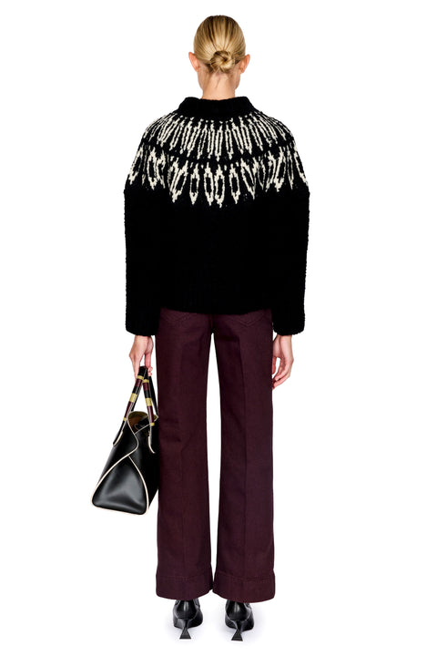A model shown from the back wearing a black knit sweater with a white geometric pattern across the shoulders, paired with dark burgundy wide-leg trousers and holding a black handbag against a plain white background.