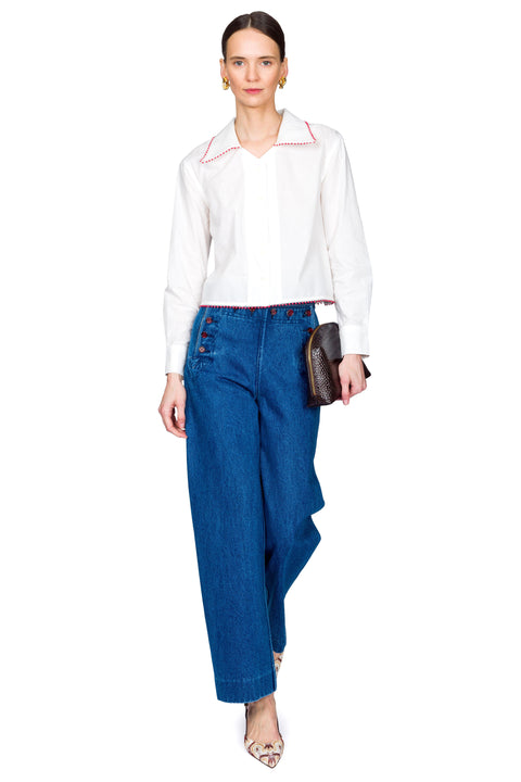 Woman wearing a white blouse with red accents and blue jeans with red buttons on a white background with one foot forward.