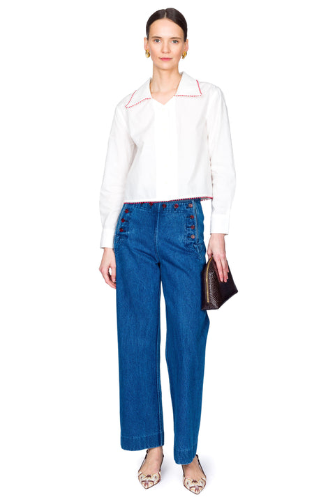Woman wearing a white blouse with red accents and blue jeans with red buttons on a white background slouching to one side.