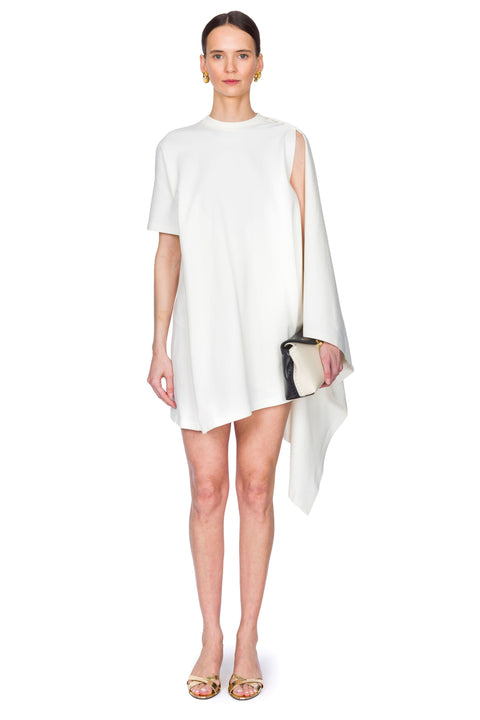 Woman wearing a white dress with a cutout design on one shoulder on a white background clutching a bag in one hand.