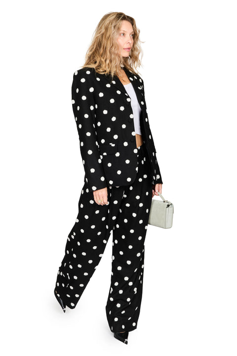 A woman modeling a black two-piece suit with white polka dots, featuring a cropped blazer and matching wide-leg trousers, styled with a white top and a small silver handbag against a plain white background.