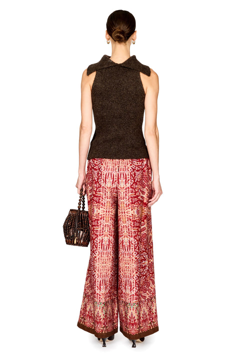 A model shown from the back wearing a sleeveless dark brown knit top and wide-leg trousers with a red, pink, and gold patterned print. She is holding a small woven brown handbag against a plain white background.