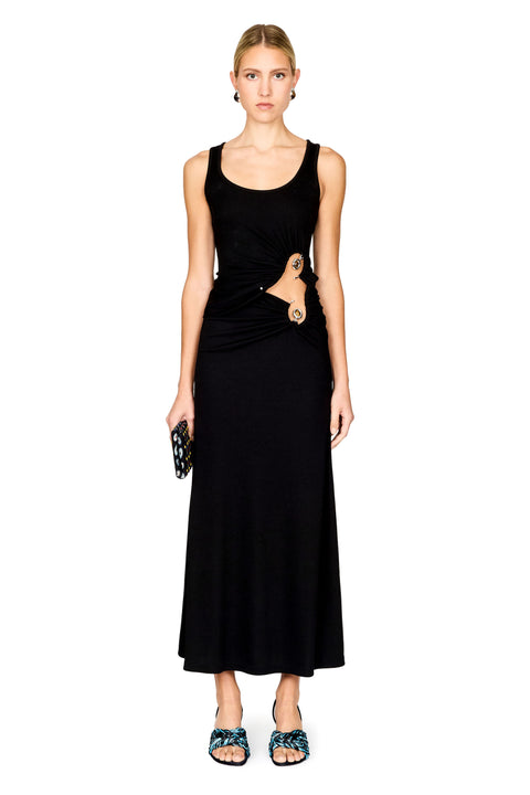 A model stands against a white background wearing a sleeveless black top and maxi skirt with a draped waist detail and a gold-toned ring accent. She is holding a small black patterned clutch and wearing blue patterned flat sandals.