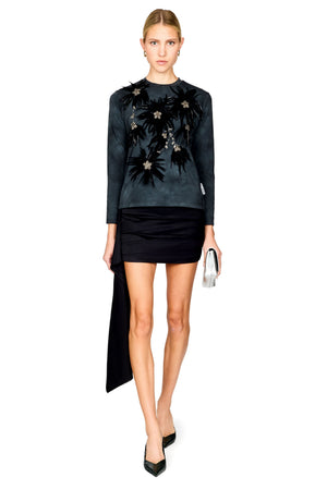 A model wearing a dark charcoal long-sleeve top with black floral embroidery and beadwork, paired with a black mini skirt and black pointed-toe heels, holding a small silver clutch against a plain white background.