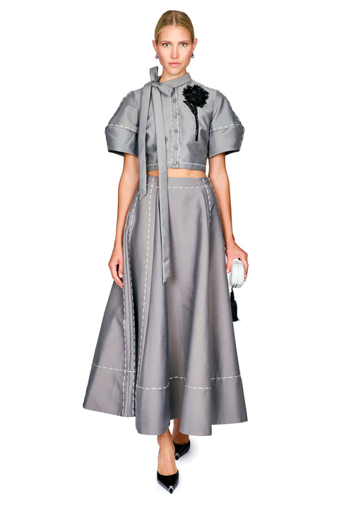 A model wearing a gray two-piece outfit consisting of a short-sleeve cropped blouse with a high neck and long tie, and a matching high-waisted flared skirt with white contrast stitching. She is holding a small white clutch against a plain white background.