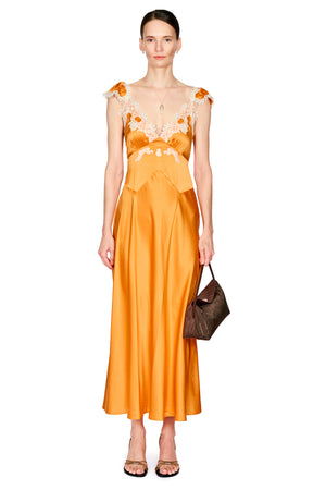 A woman wearing a vibrant orange satin maxi dress with floral lace detailing on the bodice and shoulders, holding a brown handbag against a solid white background.