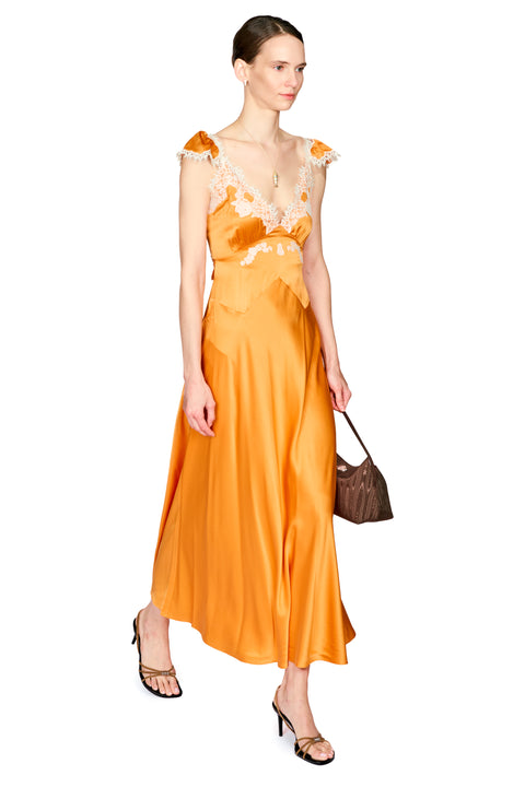 A woman modeling a bright orange satin maxi dress with white lace trim on the bodice and shoulders, paired with brown handbag and black strappy heels, against a plain white background.