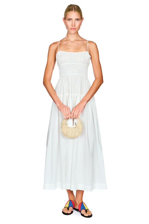 A woman modeling a white sleeveless maxi dress with thin straps, holding a round woven straw handbag and wearing colorful sandals against a plain white background.