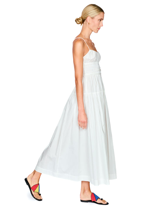 A woman modeling a white spaghetti-strap maxi dress with a gathered waist, shown in profile against a solid white background.