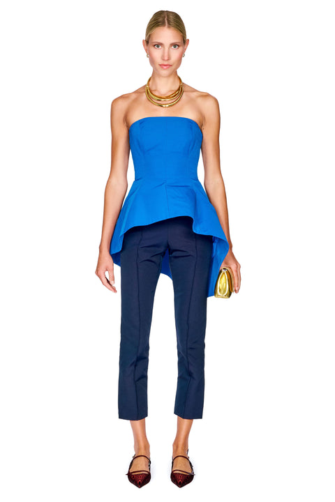A model wearing a bright blue strapless peplum top and navy blue cropped trousers, accessorized with a gold necklace and holding a gold clutch, set against a plain white background.