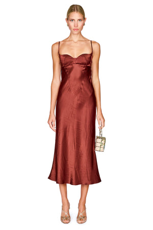 A woman modeling a satin slip midi dress in a rust brown color, styled with a small flower-patterned handbag and metallic heels, against a plain white background.