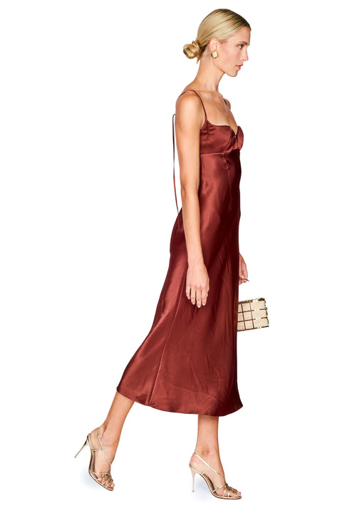A woman modeling a satin slip midi dress in a deep burgundy color, styled with nude high heels and a small beige clutch, set against a plain white background.
